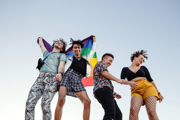 Cheerful group of queer individuals celebrating gay pride. Four members of the LGBTQ+ community dancing happily while raising the rainbow pride flag. Group of friends celebrating together outdoors.